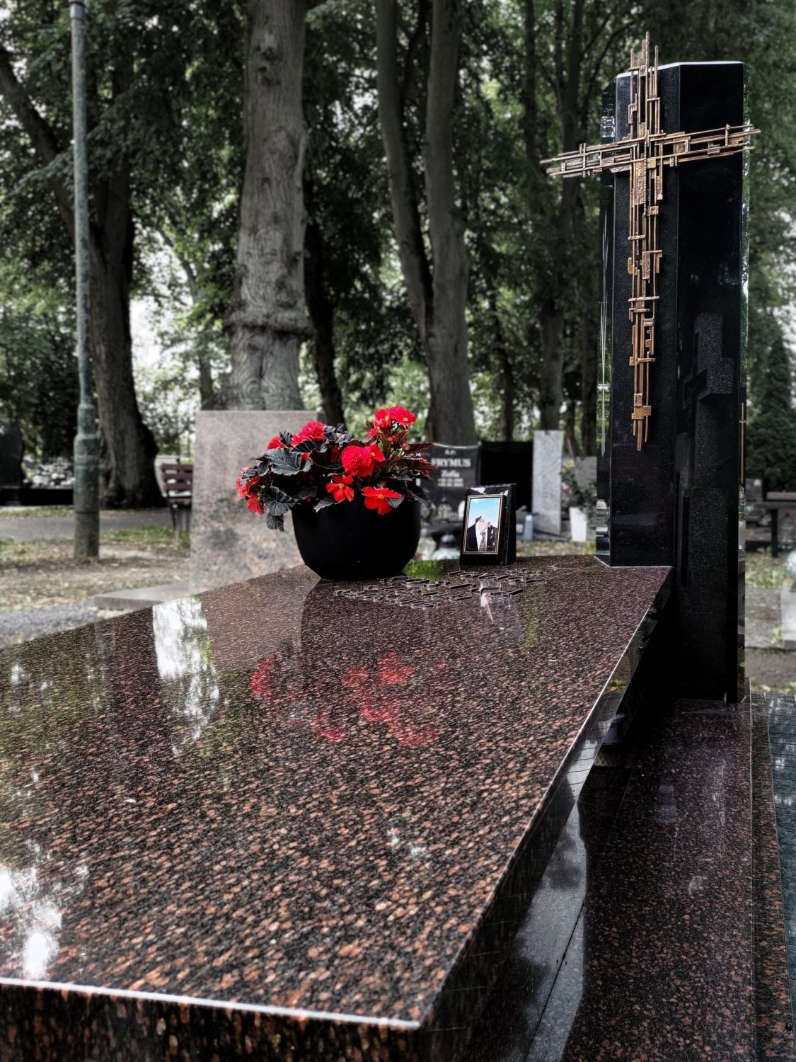 Nagrobek granitowy czerwony z czarnym słupem i miedzianym krzyżem na cmentarzu